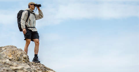 A hiker wearing a light gray zip-up hoodie, black shorts, and a black backpack stands on a rocky outcrop, looking through binoculars against a cloudy blue sky.